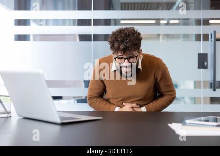 Young businessman at his office desk, hunched over laptop and holding his stomach in discomfort from a sudden abdominal pain or stomach ache while try Stock Photo