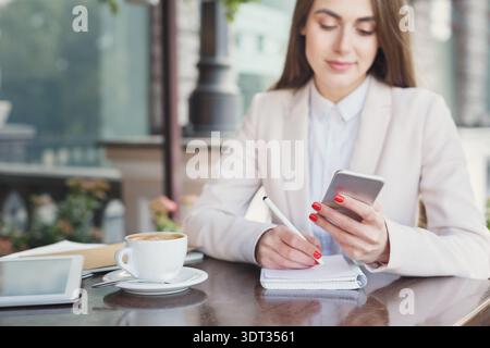 Business Woman Uses Smartphone and Takes Notes at Cafe Table During Morning Hours Stock Photo