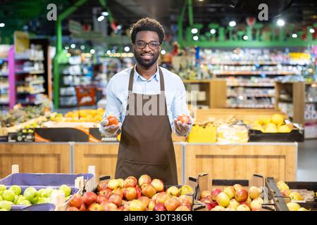 Smiling male grocer wearing an apron and gloves, standing in a supermarket produce section, holding two red apples, and looking at the camera with a friendly expression Stock Photo