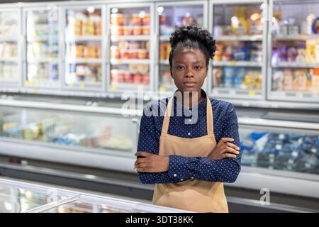 African american woman working as a supermarket employee, wearing an apron, posing confidently with crossed arms in front of the grocery store's frozen food and freezer sections Stock Photo