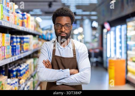 African american male worker wearing an apron and glasses standing confidently with crossed arms in a grocery store aisle, representing retail service and customer support Stock Photo