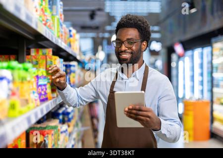 African american male worker wearing an apron and eyeglasses, checking inventory on a digital tablet while arranging products on a shelf in a modern supermarket Stock Photo