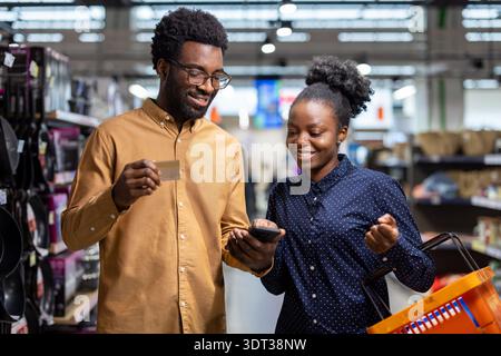 African american couple in a grocery store comparing product details and prices on a smartphone while holding a credit card and shopping basket, representing modern consumerism and digital payment Stock Photo