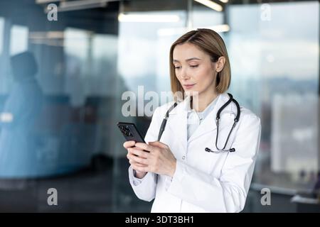 Female doctor in white coat with stethoscope holding smartphone, focused on telemedicine and digital patient care in a modern clinic, using mobile health apps for consultations Stock Photo