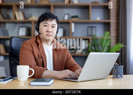 Asian man wearing a black headset sitting at a wooden desk with a laptop, typing and looking at the camera, working remotely from a modern home office with bookshelves Stock Photo