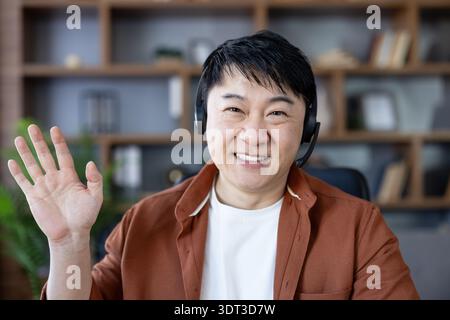 Smiling asian man in headset waving during an online video call, greeting colleagues or students from a modern home office while working or teaching remotely via webcam Stock Photo