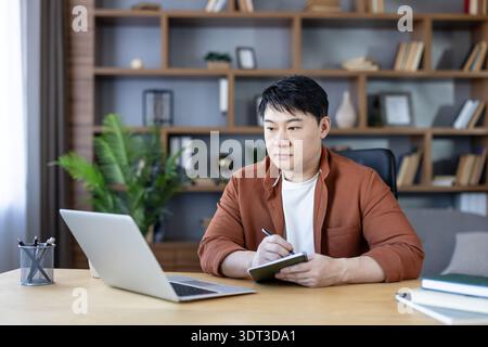 Attentive asian man focusing on a laptop screen while writing in a notebook, working or studying at a wooden desk in a modern home office with bookshelves Stock Photo