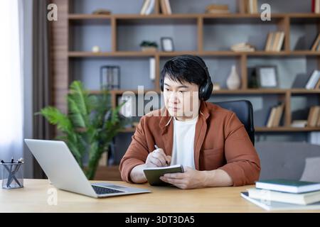 Asian man in headphones intently learning from his laptop, taking notes in a notebook while engaging in remote education or a virtual meeting from his comfortable home office Stock Photo