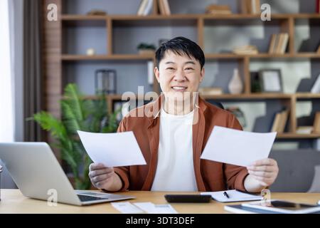 Asian man sitting at desk in home office, holding papers and looking at camera, actively managing personal finances and household budget with a laptop, feeling successful Stock Photo