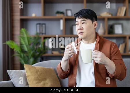 Asian man on a sofa at home holding a tissue and warm mug, looking unwell with cold flu symptoms while resting indoors during self-isolation and recovery Stock Photo