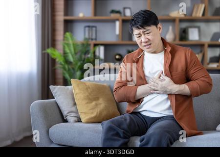Asian man experiencing severe chest pain and discomfort from a heart attack, sitting on a sofa at home and urgently clutching his chest with both hands Stock Photo