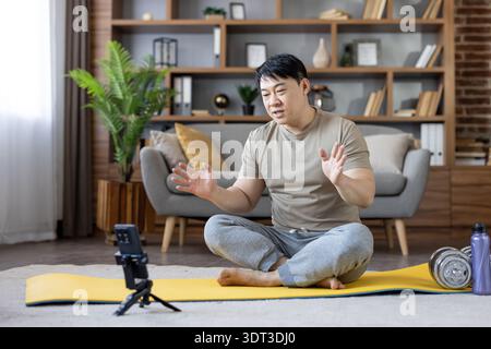 Focused asian man leading a live at-home workout from his living room, seated on a yoga mat with smartphone on a tripod, guiding virtual fitness and wellness session Stock Photo