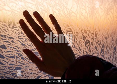 Human hand in silhouette gently pressing against a windowpane covered in beautiful, intricate frost patterns, highlighting the cold temperature as warm sunlight illuminates the natural ice formation Stock Photo