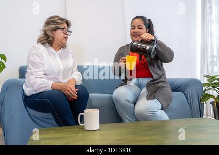 Two women relaxing on a couch, sharing coffee and having a conversation in a living room Stock Photo