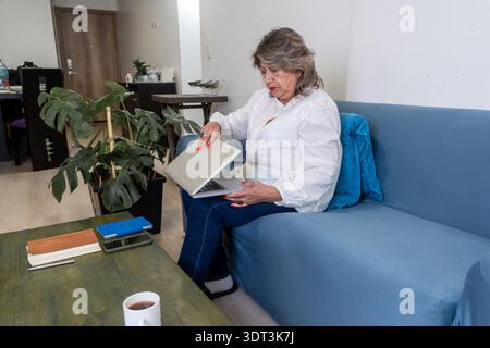 Senior woman is closing her laptop while sitting on a blue sofa in a home living room Stock Photo