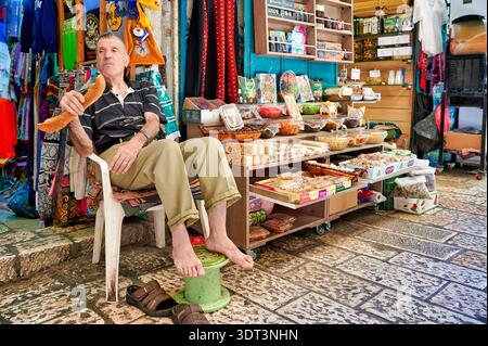 A man sits on a stool in a shop in the old city of Jerusalem. He enjoys a snack and watches customers as they browse the goods. Stock Photo