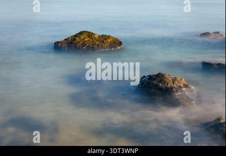 Close-up Long Exposure of Sea Waves Crashing Over Stones. A detailed long exposure shot showing the silky movement of water around dark rocks. The Stock Photo