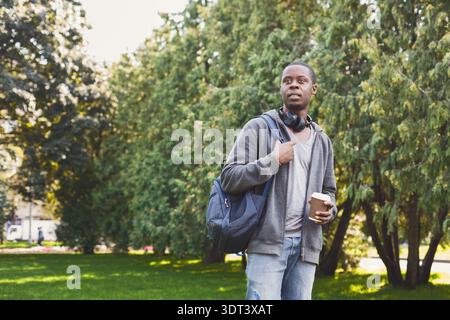 Young Man Stands in Park Holding Coffee, Wearing Headphones and Backpack on a Sunny Day Stock Photo
