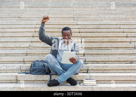 Young Man Celebrates Achievement While Sitting Outdoors With Tablet and Books on Stairs Stock Photo