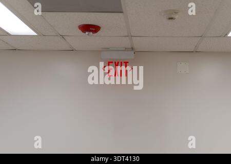 A horizontal shot of a glowing red EXIT sign mounted on a white wall below a drop ceiling. The image also captures a red fire alarm strobe and a smoke Stock Photo