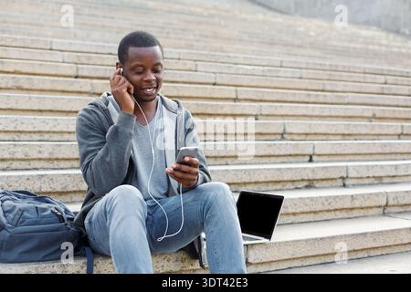 Young Man Sits on Stairs Listening to Music While Using Smartphone and Laptop Stock Photo