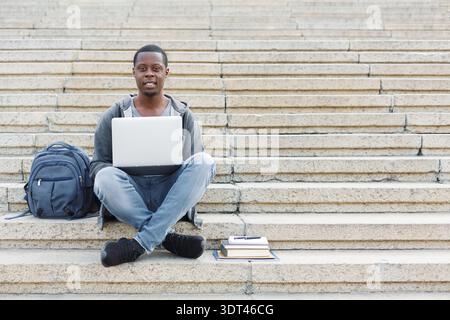 Young Man Works on Laptop While Sitting on Steps With Backpack and Books in a Public Space Stock Photo