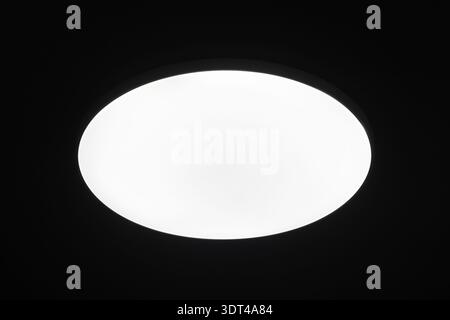 Bright circular ceiling light fixture casting an even white glow against a deep black background, highlighting minimalist modern design, ambient light Stock Photo