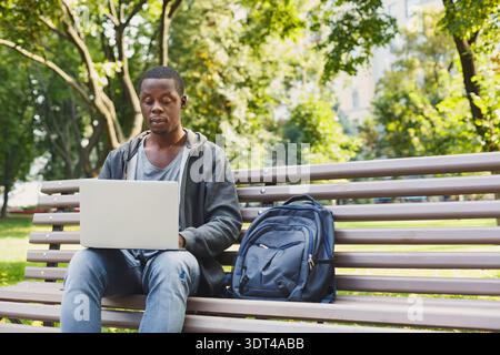 Man Working on Laptop in Park During Afternoon With Backpack Nearby on Bench Stock Photo