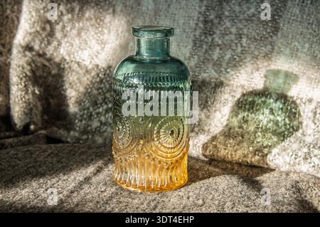 A vintage-style glass bottle with a gradient from teal to amber, featuring intricate embossed patterns and casting a distinct shadow Stock Photo