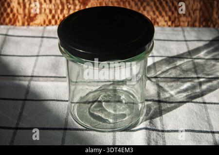 A clean, empty glass jar with a black screw-on lid rests on a white and grey checkered fabric, with dappled sunlight casting shadows Stock Photo