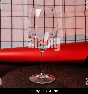 A clear, empty wine glass stands on a textured wooden plate, set against a backdrop of red and white checkered fabric and a red roll Stock Photo