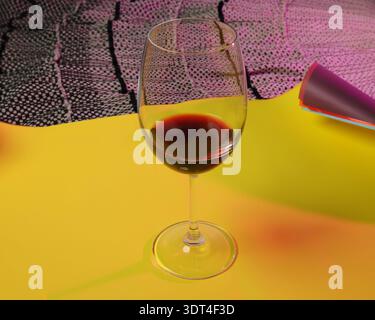 A clear wine glass filled with deep red wine is positioned on a bright yellow surface, set against a backdrop of textured fabric and rolled paper Stock Photo