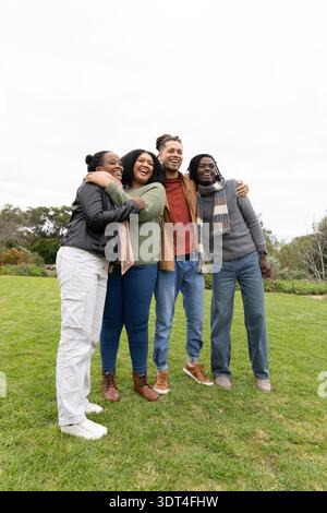 Diverse friends hugging and laughing while standing on park lawn wearing jackets and scarf Stock Photo