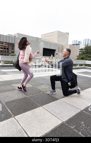 Couple on rooftop terrace with man kneeling, presenting open box woman in pink jacket raising hands Stock Photo