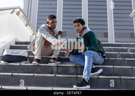 Diverse teenage male friends sitting on concrete steps in front of tower, sharing tablet and coffee Stock Photo