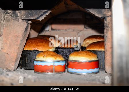 A horizontal shot showing several loaves of golden brown bread baking inside a rustic brick oven. The bread is in various metal tins, including tradit Stock Photo