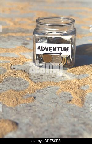 Collection of Coins in a Jar Labeled Adventure on a Sandy Surface and Sunlight Reflecting Stock Photo