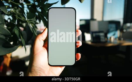 Smartphone mockup empty screen modern office interior plants. Hand holding device against cozy workspace, lush green leaves foreground, warm sunlight Stock Photo