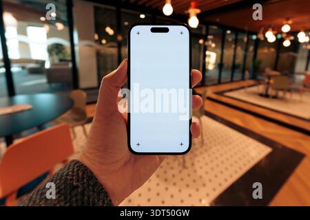 Smartphone mockup empty screen corporate cafeteria lounge. Hand holding modern mobile device within stylish company canteen, glass walls, wooden ceili Stock Photo