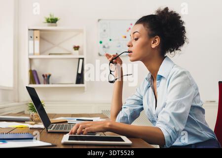 Woman Working at a Desk While Thinking and Using a Laptop With Glasses in Hand Stock Photo