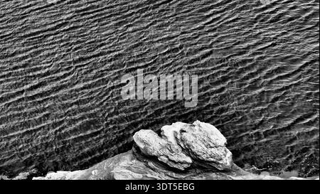 Black and white minimalist coastal scene featuring layered rock formations in the foreground and wind-rippled seawater creating textured patterns acro Stock Photo