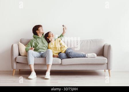 Split system. Happy young Asian couple with remote relaxing under air conditioner, sitting on couch at home, mockup Stock Photo
