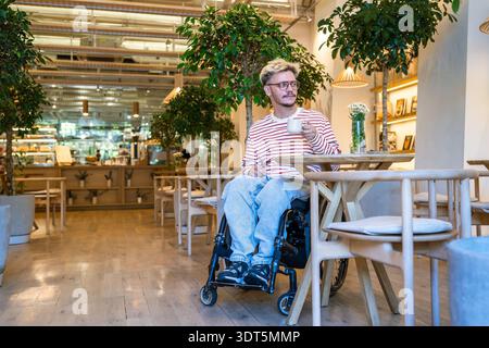 Relaxed positive disabled man in wheelchair drinking coffee in trendy cafe, staying socially active. Stock Photo