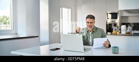 Senior mature man looking at laptop checking bill working at home kitchen table. Stock Photo