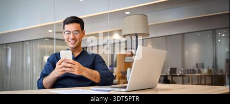 Smiling busy Hispanic business man sitting in office holding mobile phone. Stock Photo