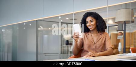 Young happy African American business woman using cell phone sitting in office. Stock Photo