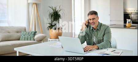 Happy older mature man talking on phone using laptop at home. Stock Photo
