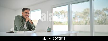 Happy older mature man using laptop computer at home table. Stock Photo