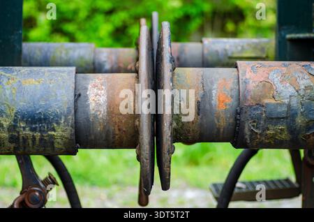 Close-up of rusty industrial shaft with metal coupling wheels on old machinery, weathered paint, outdoors with green blurred background, gritty textur Stock Photo