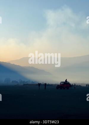 Jeep and small figures crossing a misty volcanic plain beneath smoky clouds and soft morning light Stock Photo